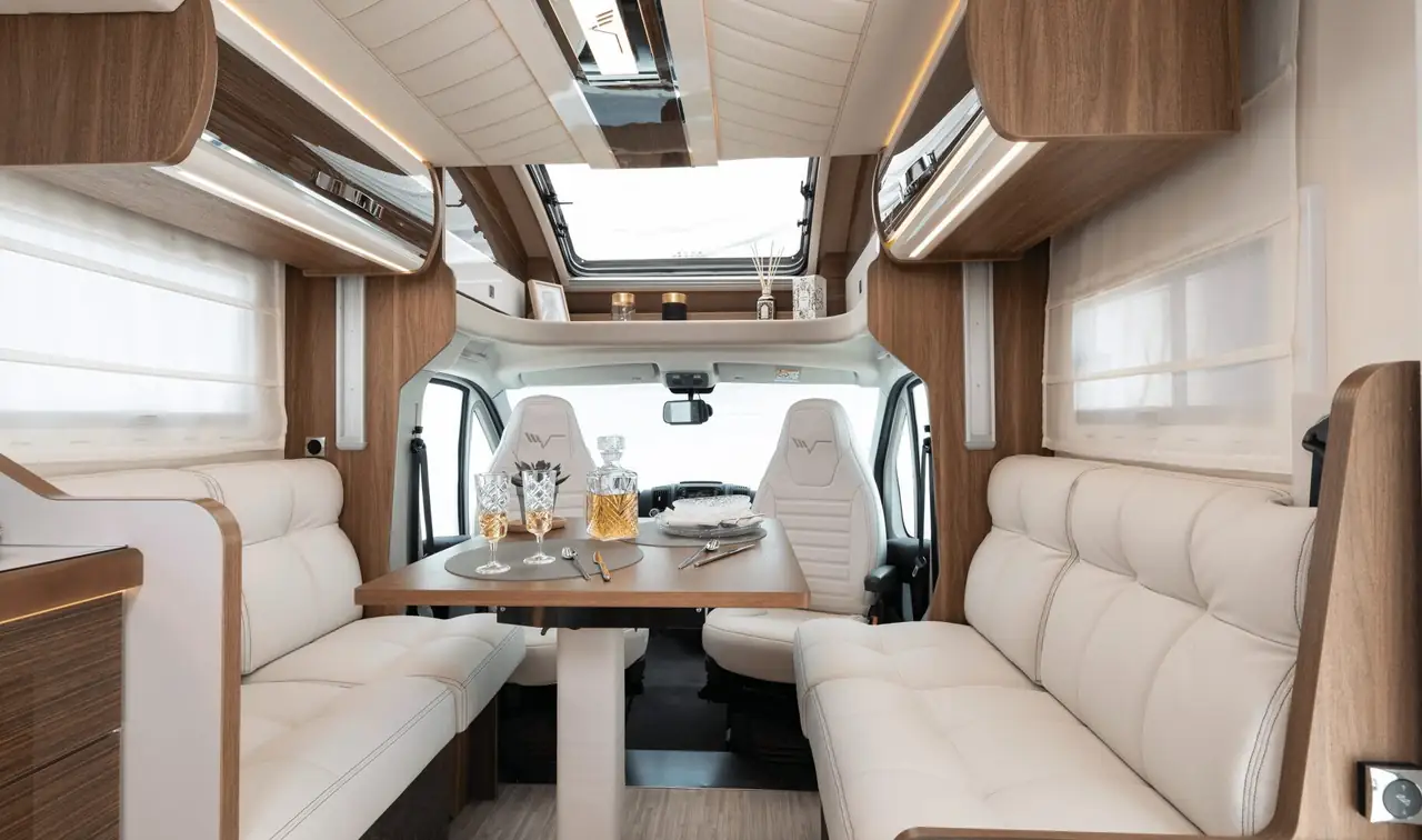 La dinette di un camper: divani in pelle chiara, il tavolo in legno apparecchiato, i pensili e le pareti rivestite.